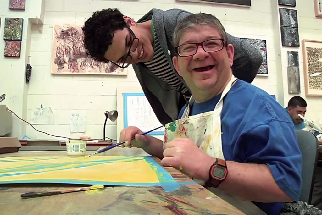 A man wearing glasses and a paint-splattered apron smiles while painting at a table; another person leans over and looks at his artwork in an art studio.