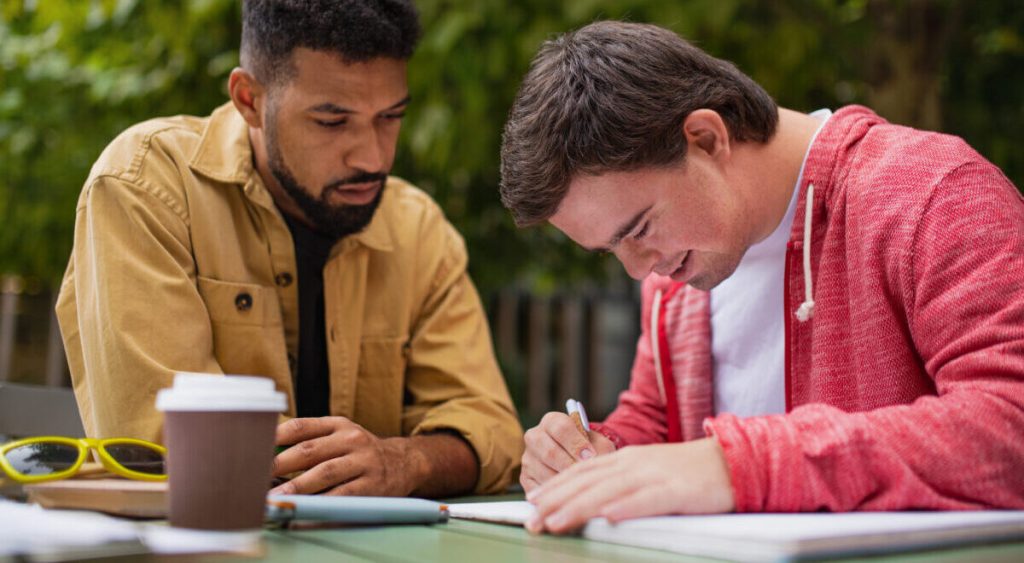A young man with Down syndrome with his mentoring friend sitting outdoors in cafe and studying.